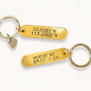 May include: Two gold-colored keychains with rectangular tags. One tag has coordinates, the other reads "WHERE WE SAID I DO." Each keychain has a round ring and a small heart charm.