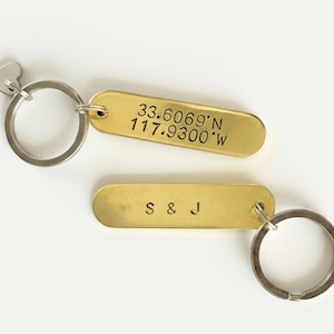 May include: Two brass keychains with silver key rings. One keychain has the coordinates 33.6069°N 117.9300°W engraved on it. The other keychain has the initials "S & J" engraved on it. The keychains are on a white background.