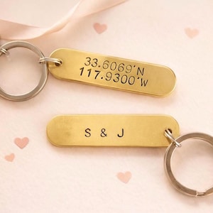 May include: Two brass keychains with silver key rings. One keychain has the coordinates 33.6069°N 117.9300°W engraved on it. The other keychain has the letters "S & J" engraved on it. Pink heart-shaped confetti is scattered around the keychains.