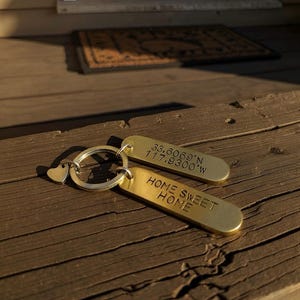 May include: Two brass keychains with a heart-shaped charm and a key ring. One keychain has the coordinates 33.6069°N 117.9300°W, and the other says "HOME SWEET HOME". The keychains are on a wooden surface.