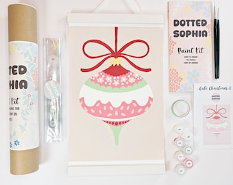 Puede incluir: Un kit de pintura por números con un lienzo colgante que muestra una ilustración de un adorno navideño rojo, rosa y verde. Incluye un tubo de cartón, un folleto guía Dotted Sophia Paint Kit, dos pinceles, una cinta adhesiva y botes de pintura numerados.