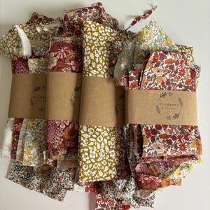 May include: Bundles of floral fabric in various patterns and colors, including red, yellow, and pink. Each bundle is wrapped with a brown paper band that has the text "wildseed stitches". The fabric appears to be cotton or a similar material.