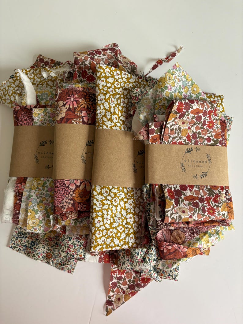 May include: Bundles of floral fabric in various patterns and colors, including red, yellow, and pink. Each bundle is wrapped with a brown paper band that has the text "wildseed stitches". The fabric appears to be cotton or a similar material.