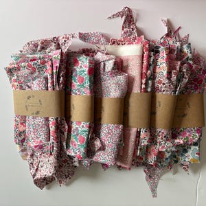 May include: A collection of floral fabric bundles in shades of pink, red, and white, wrapped with brown paper bands. The fabric features various floral patterns, including small flowers and strawberries. The bundles are arranged in a neat stack.