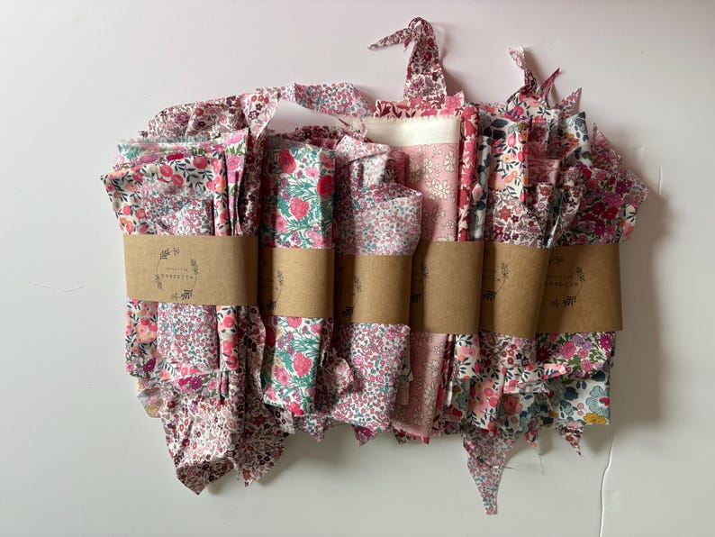 May include: A collection of floral fabric bundles in shades of pink, red, and white, wrapped with brown paper bands. The fabric features various floral patterns, including small flowers and strawberries. The bundles are arranged in a neat stack.