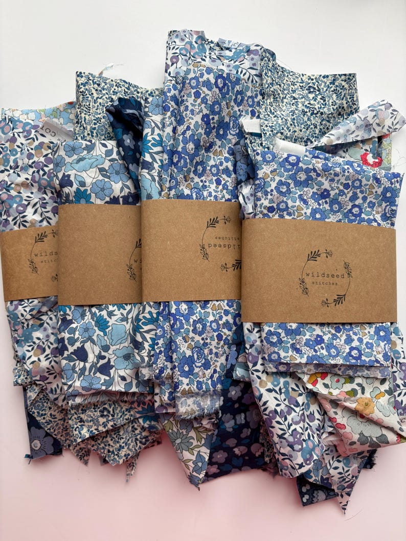 May include: Several bundles of floral fabric in shades of blue, white, and purple are displayed. Each bundle is wrapped with a brown paper band featuring the text "wildseed stitches". The fabric patterns include small flowers and leaves.