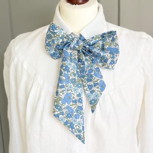 May include: A white long-sleeved button-down shirt featuring a large, tied neck bow accessory. The bow is made of fabric with a dense floral pattern in shades of light blue, medium blue, and sage green against a white background.