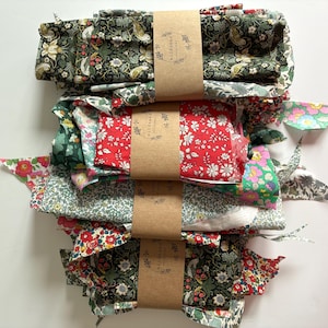 May include: A stack of fabric bundles in various floral patterns, including red, green, and white. Each bundle is wrapped with brown paper and tied with a matching label. The fabric appears to be cotton or a similar lightweight material, suitable for quilting or crafting.