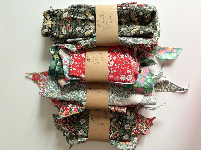 May include: A stack of fabric bundles in various floral patterns, including red, green, and white. Each bundle is wrapped with brown paper and tied with a matching label. The fabric appears to be cotton or a similar lightweight material, suitable for quilting or crafting.