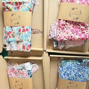 May include: Four cardboard boxes filled with floral fabric bundles. Each bundle is wrapped with a brown paper band featuring a logo. The fabrics display various floral patterns in shades of pink, blue, and red. The boxes are arranged in a grid.