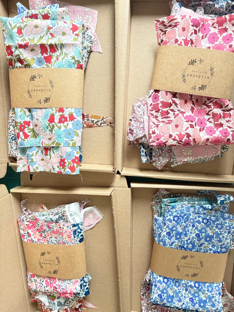 May include: Four cardboard boxes filled with floral fabric bundles. Each bundle is wrapped with a brown paper band featuring a logo. The fabrics display various floral patterns in shades of pink, blue, and red. The boxes are arranged in a grid.