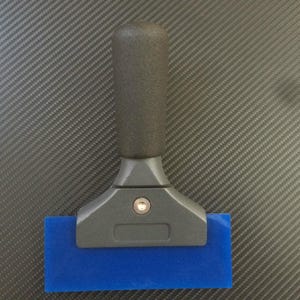 Puede incluir: Una espátula azul con mango negro y base gris. La espátula tiene una hoja azul rectangular y está diseñada para aplicar vinilo o tintar ventanas. El mango es texturizado para un mejor agarre.