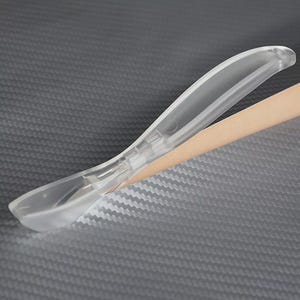 Puede incluir: Una cuchara de plástico transparente con un mango curvo. La cuchara tiene un cuenco pequeño de forma ovalada y está diseñada para recoger. La cuchara está colocada sobre un fondo gris texturizado con un objeto de color beige.