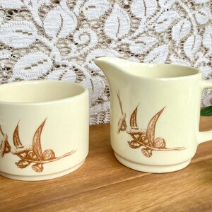May include: Cream-colored ceramic creamer and sugar bowl set. Each piece features a brown floral design. The creamer has a handle and spout. The sugar bowl is cylindrical. The set is on a wooden surface.