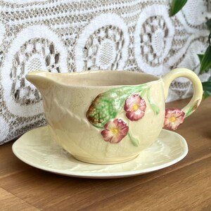 May include: A cream-coloured ceramic gravy boat and saucer set. The gravy boat has a spout and handle, decorated with raised floral and leaf designs in pink, green, and cream. The saucer has a matching embossed leaf pattern.