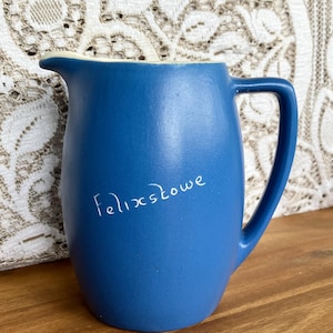 May include: A blue ceramic pitcher with a curved handle and a spout. The pitcher has the word "Felixstowe" written on the side in white script. The interior rim is a cream color.