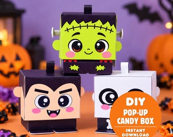 May include: Three square paper candy boxes featuring cartoon characters: a green Frankenstein monster, a black-haired vampire, and a white ghost. Each box has a pull tab on top and displays the text DIY POP-UP CANDY BOX INSTANT DOWNLOAD.