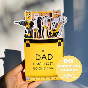 May include: A yellow DIY chocolate toolbox card with the text "IF DAD CAN'T FIX IT, NO ONE CAN!" and illustrations of tools. The card includes a drill, saw, wrench, and other tools. The card is held by a hand.