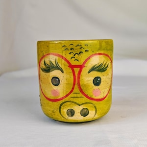 Puede incluir: Una maceta de cerámica amarilla pintada a mano con un diseño de cara caprichoso. La cara presenta gafas con montura roja, ojos grandes, mejillas rosadas y un hocico de cerdo. La maceta está decorada con pequeños detalles negros.