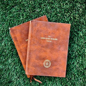 May include: Two brown leather journals with the text "The GOOD HABIT BUILDER Journal" and a compass emblem. The journals are lying on a bed of green grass.