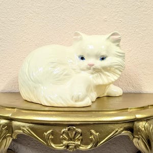 May include: A white ceramic cat figurine with blue eyes rests on a gold-painted, ornate wall shelf. The shelf has a semi-circular top and decorative, curved supports. The cat is posed in a relaxed position.
