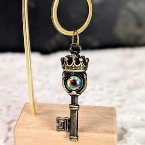 May include: Antique bronze key-shaped keychain with a crown and an eye-like design. The key is suspended from a gold-colored ring. The eye design features red, blue, and green colors. The keychain is displayed on a small wooden block.