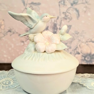 May include: A ceramic trinket box with a lid, featuring a light blue bird perched above a pink flower. The box is a soft, creamy white color, with a matching lid. The background has a floral pattern.