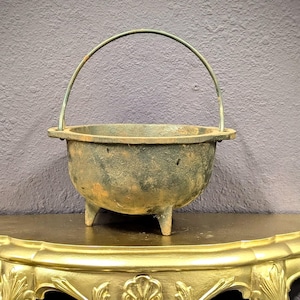 May include: An antique cast iron cauldron with a handle, resting on a gold-painted decorative shelf. The cauldron has three small legs and a weathered appearance. The shelf features ornate carvings and a semi-circular shape.