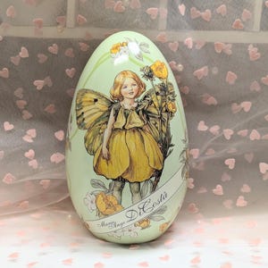 May include: A decorative egg-shaped container with a light green background. The egg features a detailed illustration of a fairy in a yellow dress and butterfly wings, holding yellow flowers. The name "Marie Ange Di Costa" is written on the egg.