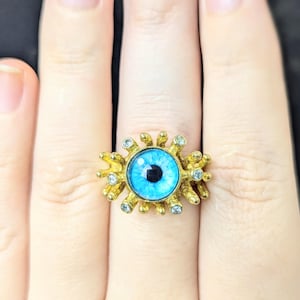 May include: A gold-colored ring featuring a large, detailed eye design. The eye has a bright blue iris with a black pupil, surrounded by a gold setting with small, clear accent stones. The ring is on a finger.