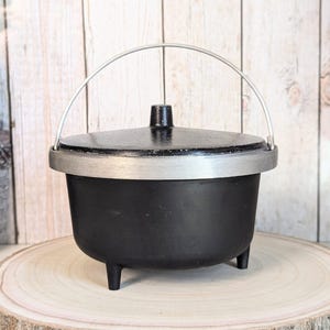 May include: A black cast iron cauldron with a lid and a silver handle. The cauldron has three small legs and a silver rim. The lid has a small black knob. The cauldron is sitting on a wooden slice.