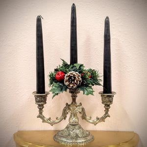 May include: An ornate, antique brass candelabra with three black taper candles. The center of the candelabra is decorated with faux greenery, red berries, and a pinecone. The candle holders have a detailed, vintage design.