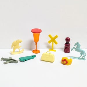 May include: A collection of small, colorful plastic toys. Includes a yellow rocking horse, an orange goblet, a yellow railroad crossing sign, a red chess piece, a blue horse, a green airplane, a green boat, a yellow suitcase, and a yellow ring.