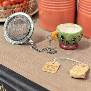 May include: A stainless steel tea infuser with a chain is next to a small, green teacup with an orange handle and red base. A tea bag and a tag that reads "STEEPED WITH KINDNESS" are also present. The items are on a brown surface.