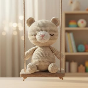 Peut inclure: Un ours en peluche crocheté beige, assis sur une balançoire en bois. L'ours a un nez rose et les yeux fermés. La balançoire est suspendue par une corde beige. L'arrière-plan est flou, avec une étagère visible.