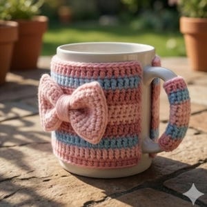 Peut inclure: Une tasse en céramique blanche avec une housse crochetée. La housse est rayée rose, bleu clair et rose avec un grand nœud. La poignée est également recouverte du même matériau crocheté. La tasse est posée sur une surface en pierre.