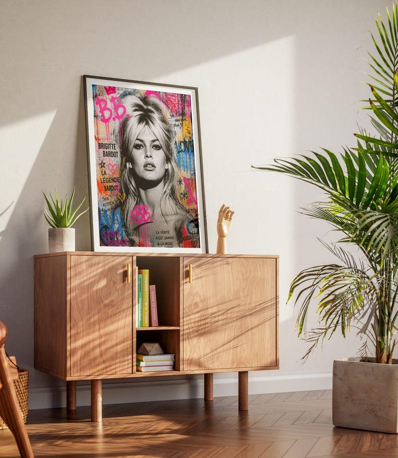Puede incluir: Una impresi&oacute;n art&iacute;stica enmarcada con una imagen en blanco y negro de Brigitte Bardot con acentos coloridos de estilo graffiti. La obra de arte incluye el texto "Brigitte Bardot" y otras frases francesas. La impresi&oacute;n se exhibe en un mueble de madera con una planta y objetos decorativos.