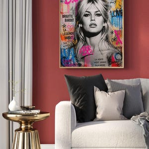 Puede incluir: Una obra de arte enmarcada con un retrato en blanco y negro de Brigitte Bardot, con textos y acentos coloridos de estilo graffiti. El texto incluye "BB", "Brigitte Bardot" y "La L&eacute;gende Bardot". La obra de arte cuelga de una pared roja.