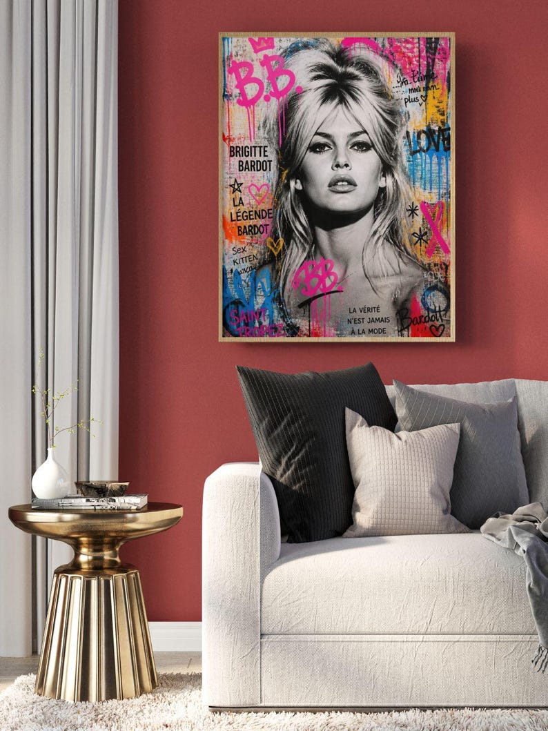 Puede incluir: Una obra de arte enmarcada con un retrato en blanco y negro de Brigitte Bardot, con textos y acentos coloridos de estilo graffiti. El texto incluye "BB", "Brigitte Bardot" y "La L&eacute;gende Bardot". La obra de arte cuelga de una pared roja.