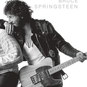 May include: Black and white image of Bruce Springsteen. He is wearing a black leather jacket, a white undershirt, and a necklace. He is holding a wooden electric guitar. The text "BRUCE SPRINGSTEEN" is at the top of the image.