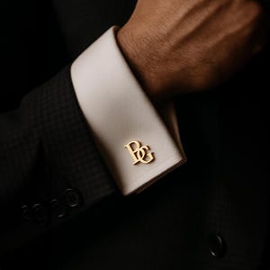 Może przedstawiać: Złote spinki do mankietów ze splecionymi inicjałami "BG" na białym mankiecie koszuli. Mankiety są częścią ciemnoszarej marynarki. Obraz jest zbliżeniem, podkreślającym szczegóły spinek do mankietów i garnituru.