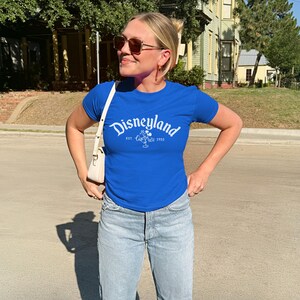 Op de afbeelding: Een koningsblauw t-shirt met het woord "Disneyland" in witte letters over de borst. Het shirt heeft ook een afbeelding van Mickey Mouse en het jaar "1955". Het shirt wordt gecombineerd met lichtblauwe jeans.