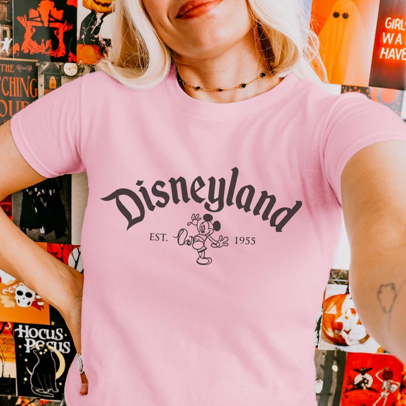 Op de afbeelding: Een lichtroze T-shirt met het woord "Disneyland" in een sierlijk lettertype, vergezeld van "EST. 1955" en een Mickey Mouse-illustratie. Het T-shirt heeft een ronde hals.