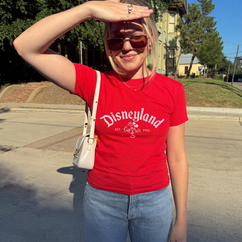 Op de afbeelding: Een rood T-shirt met het Disneyland-logo in wit, met de tekst "Disneyland Est. 1955" en een Mickey Mouse-afbeelding. Het shirt wordt gecombineerd met lichtblauwe jeans en een witte handtas.