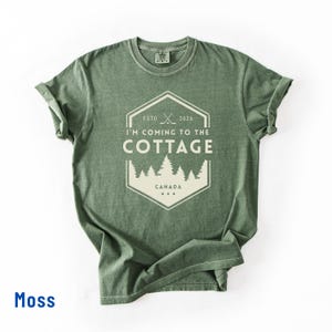 May include: Moss green t-shirt with a white graphic that reads "I'M COMING TO THE COTTAGE" with a forest and the word "CANADA" below. The shirt has a crew neck and rolled-up sleeves.