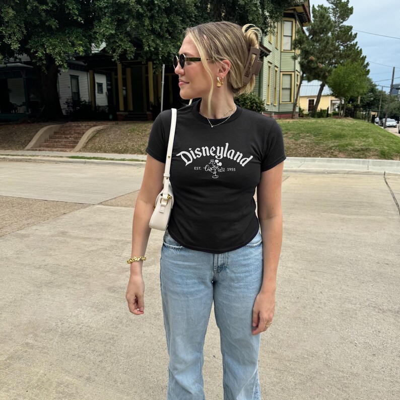 Op de afbeelding: Een persoon draagt een zwart t-shirt met het woord "Disneyland" in witte letters. Het shirt heeft ook een Mickey Mouse-afbeelding en de tekst "EST. 1955". De persoon draagt een lichtblauwe jeans, een zonnebril en een witte handtas.