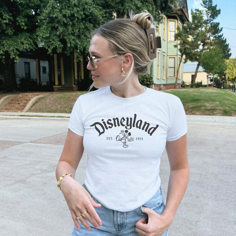 Op de afbeelding: Wit T-shirt met het woord "Disneyland" in een gebogen, zwarte letter, met een Mickey Mouse-afbeelding en de tekst "EST. 1955". Het shirt heeft korte mouwen.