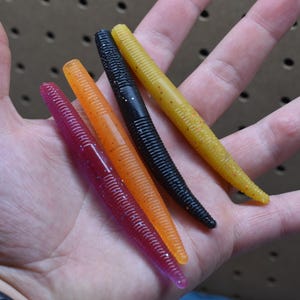 May include: Four soft plastic fishing lures in a cylindrical shape with ribbed textures. The lures are colored translucent pink, translucent orange with glitter, solid black, and translucent yellow with glitter, all arranged in a row.