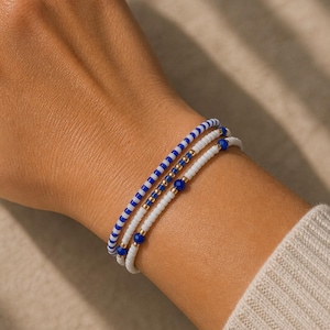 Può includere: Tre braccialetti di perline bianche, blu e oro. Uno presenta perline blu e bianche alternate, un altro ha perline bianche con dettagli dorati e il terzo ha perline blu e oro. I braccialetti sono sovrapposti su un polso.
