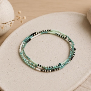 Peut inclure: Un bracelet composé de plusieurs rangs de petites perles en verre dans des tons turquoise, vert menthe, noir et blanc, alternées avec des perles intercalaires métalliques de couleur argentée, disposées en cercle sur une surface plane.